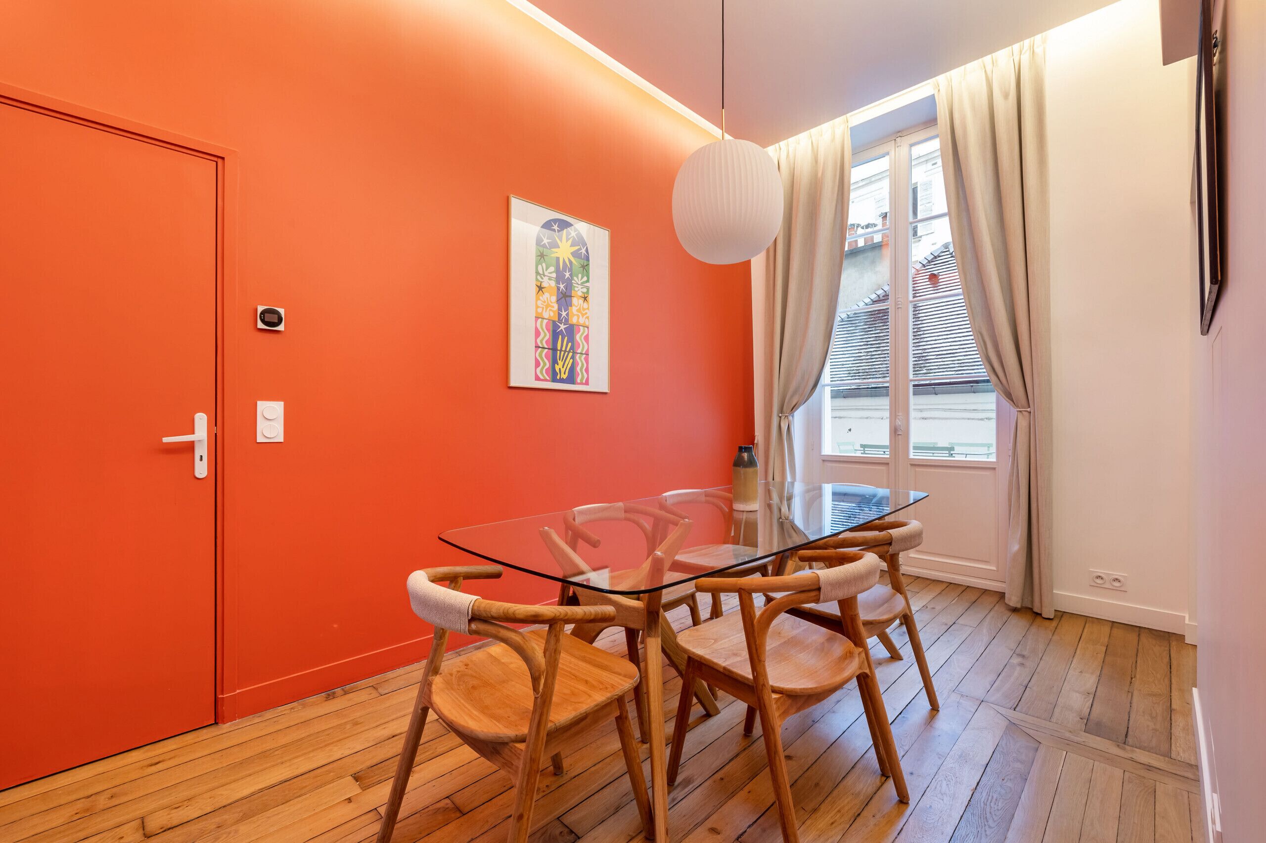Een kamer met een oranje wand, een glazen tafel, houten stoelen, een witte hanglamp en een raam met gordijnen.