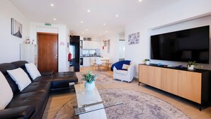 Un salon moderne avec un canapé, une table basse, un téléviseur et un coin cuisine à l’arrière-plan.
