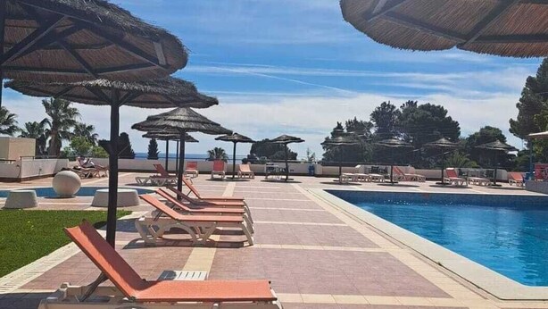 Un espace aménagé au bord de la piscine, avec des chaises longues et des parasols en chaume, un ciel dégagé et une vue sur la mer.
