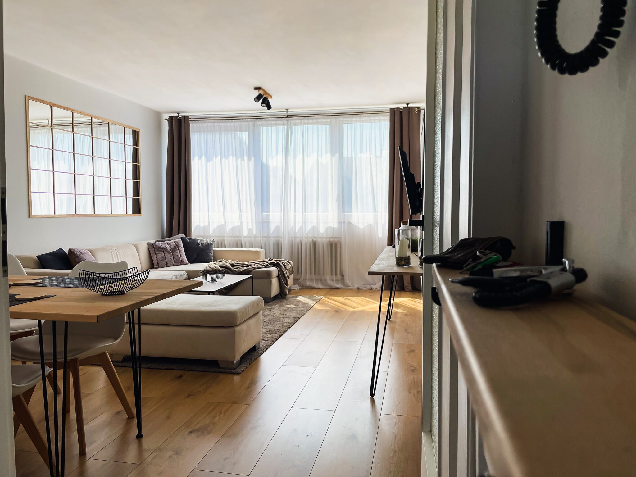 Ein modernes Wohnzimmer mit Sofa, Couchtisch und Esstisch. Ein großes Fenster mit Vorhängen, ein Heizkörper und eine Deckenleuchte sind zu sehen.