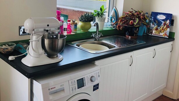 A kitchen with a built-in washing machine, a window with a view of trees, and various kitchen items on the counter. - Family-sized house 5 minutes away from the beach (Swanage)