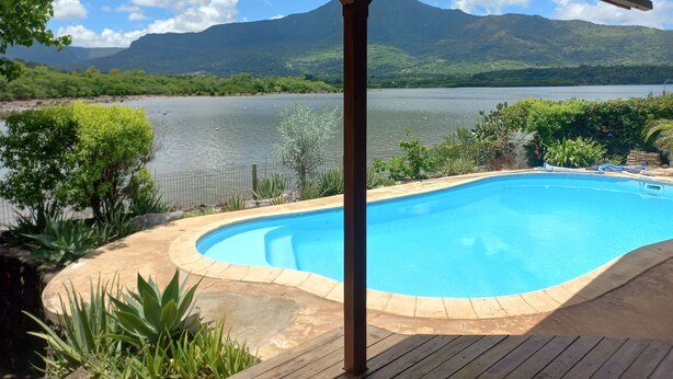 A swimming pool with a wooden deck, surrounded by lush greenery and a view of a lake with mountains in the background.