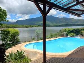 A swimming pool with a wooden deck, surrounded by lush greenery and a view of a lake with mountains in the background. - Holiday Home on Ilot Fortier Sea Shore  (Petite Rivière Noire)