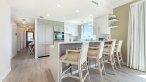 A modern kitchen with white cabinetry, a central island with bar stools, and a view of the sea through the window. - Beachcomber 24 by Grand Cayman Villas (7 Mile Beach)