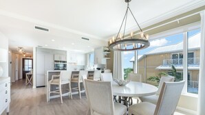 A spacious kitchen with white cabinets, a dining area with a round table and chairs, and large windows offering a view of the outside. - Beachcomber 24 by Grand Cayman Villas (7 Mile Beach)