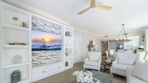 A modern living room with a large TV, white furniture, and a wooden ceiling fan. - Beachcomber 24 by Grand Cayman Villas (7 Mile Beach)