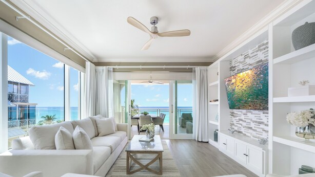 A spacious living room with a large window overlooking the ocean, a ceiling fan, and a modern art piece on the wall. - Beachcomber 24 by Grand Cayman Villas (7 Mile Beach)