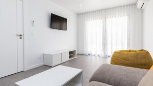 Una sala de estar moderna con un sofá blanco, una mesa de centro blanca, un sillón color mostaza, un televisor de pantalla plana montado en la pared y un ventanal con cortinas translúcidas.