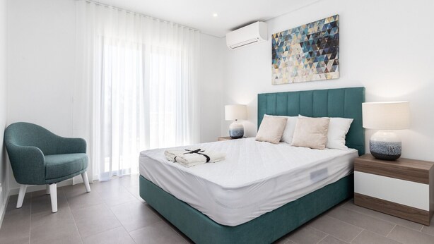 Una habitación de hotel moderna con una cama color turquesa, cortinas blancas, un sillón color turquesa y una obra de arte geométrica en la pared.