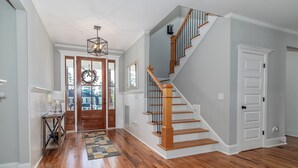 A spacious hallway with hardwood flooring, a wooden staircase with a glass railing, and a door with a decorative wreath. - Perfect Masters Rental in Golf Course Community - Bartram Trail CC: Augusta, GA  (Evans)