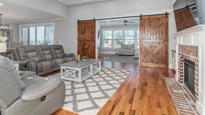 A spacious living room with a grey leather sofa, a white coffee table, and a patterned rug. A wooden door with a cross-brace design leads to a balcony with a view of the outdoors. - Perfect Masters Rental in Golf Course Community - Bartram Trail CC: Augusta, GA  (Evans)