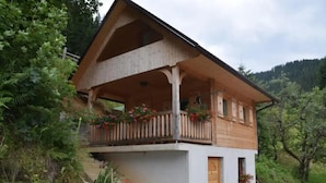 A wooden chalet with a balcony, surrounded by greenery and a mountainous backdrop.