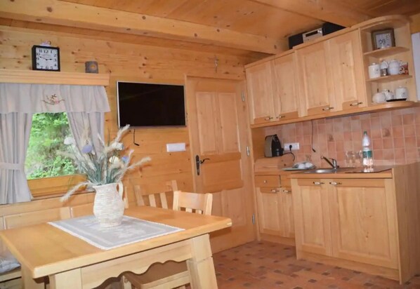 Interior de una cabaña de madera con una mesa de comedor, sillas, un jarrón con flores, un reloj, un televisor, armarios de madera y un salpicadero de azulejos.