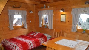 A cozy cabin with a wooden interior, a bed with red bedding, a dining table, and two windows with curtains.