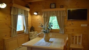 A wooden cabin interior with a dining table, chairs, a vase with flowers, a clock, and a TV on the wall.