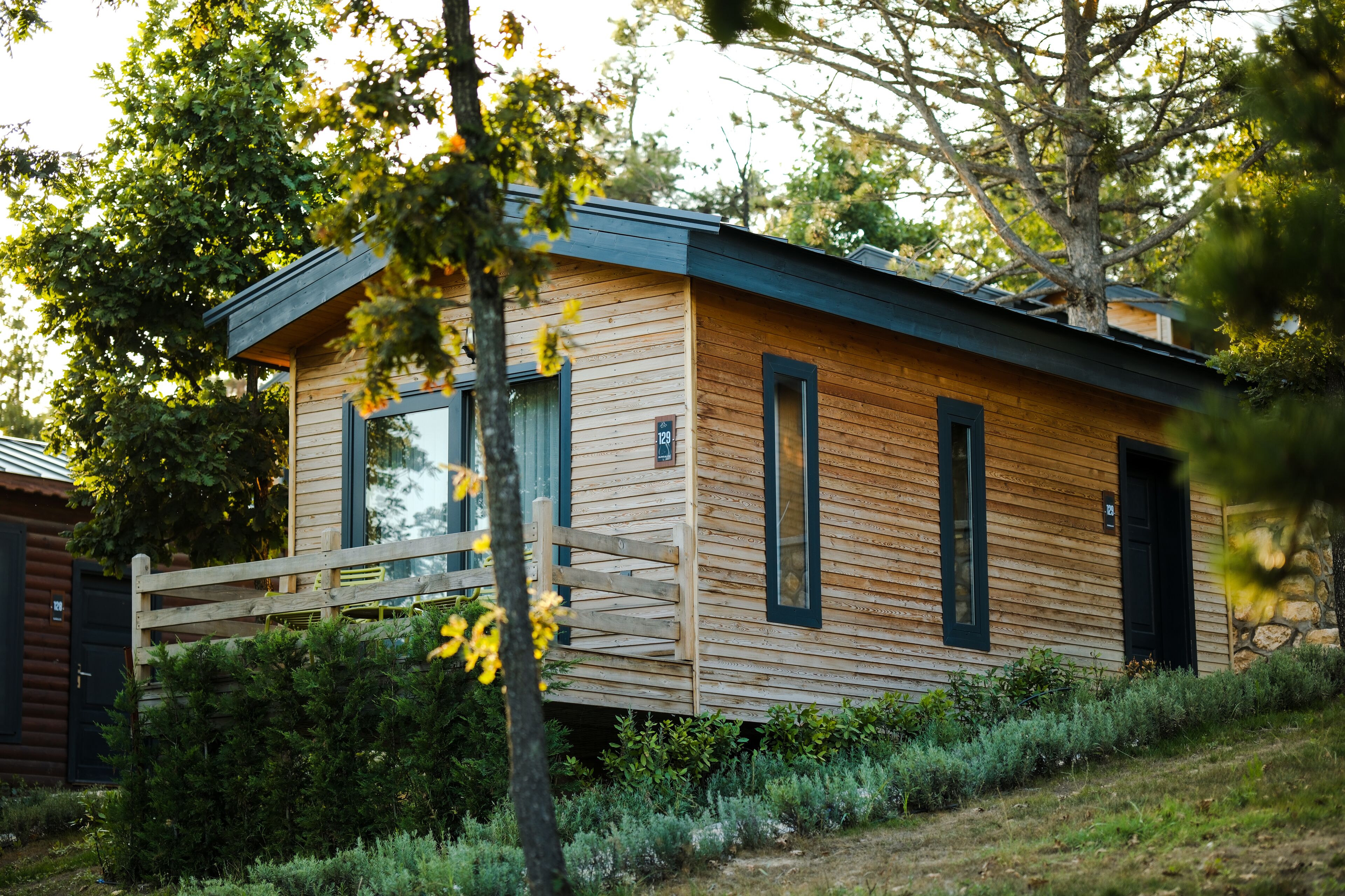 A modern wooden cabin with a deck, surrounded by greenery.