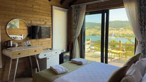 Ein Zimmer in einer Holzhütte mit Bett, Schreibtisch, Spiegel, Fernseher und Blick auf einen See und Hügel.
