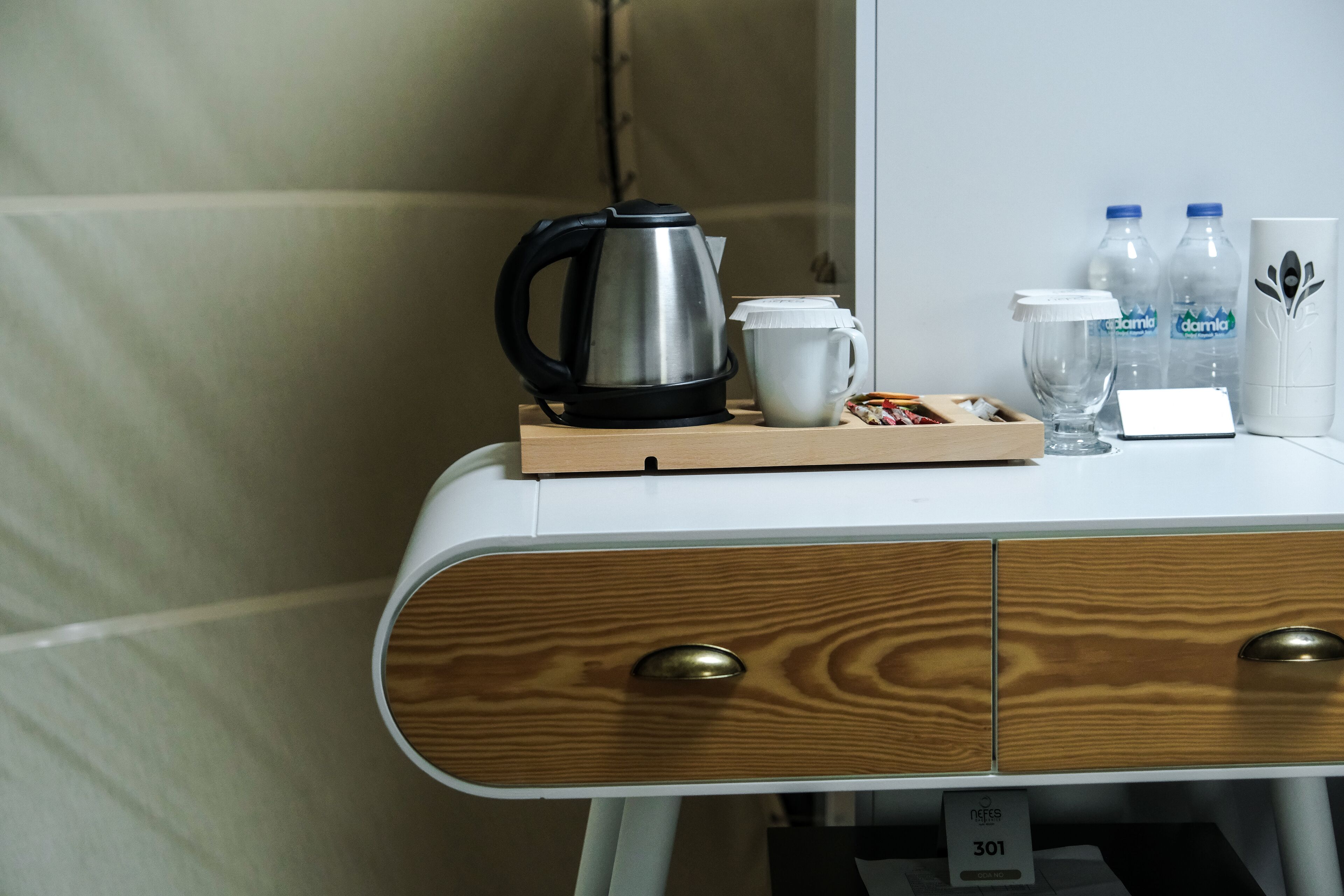 A hotel room with a wooden drawer unit, a kettle, a cup, and two water bottles on a small table.