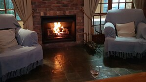 Two armchairs with fringed covers facing a fireplace with a visible fire. - - Country getaway in Peaceful setting yet minutes from Town (Gqeberha)