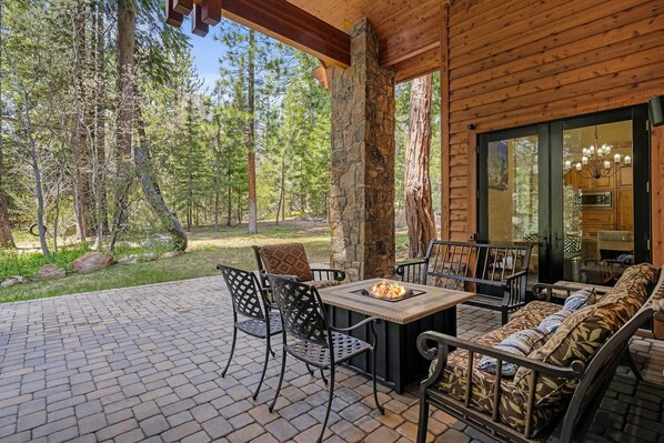 A covered patio with a stone fireplace, outdoor seating, and a view of a forest. - 5BR Incline Village Retreat | Hot Tub & Mountain Views (Incline Village)