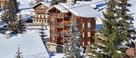 A multi-story wooden building with balconies, surrounded by snow-covered trees and a ski slope in the background.