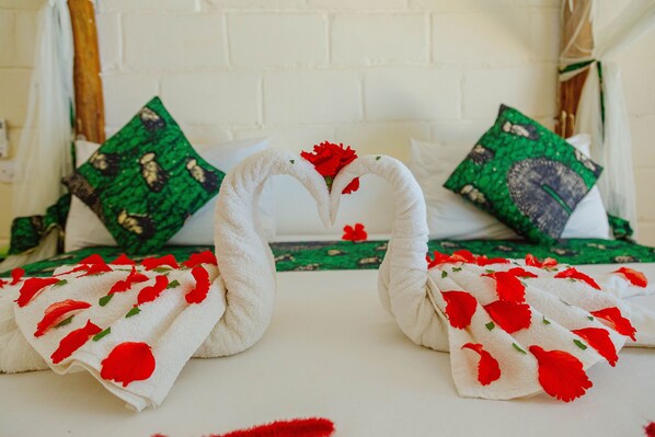 A bed with a white sheet and two swan-shaped towels with red flowers. There are green pillows with a leaf pattern. - Tropicana Garden (Kendwa)