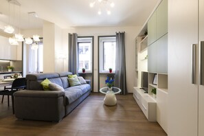 Een moderne woonkamer met een grijze bank, een glazen salontafel en een keuken met witte kasten en een koelkast.