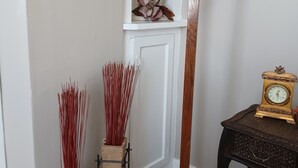 A corner of a room with a white wall, a wooden floor, a decorative wall piece, a tall white vase, a woven hat on a stand, a small table with a clock, and two tall vases with red sticks.