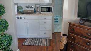 A compact kitchen with white cabinets, a microwave, and a coffee maker. There's a wooden dresser and a plant in the foreground.