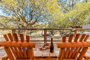 Une terrasse en bois avec deux chaises, une table sur laquelle se trouvent une bouteille et des verres, et une vue sur des arbres et une clôture.