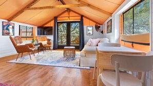 Um loft espaçoso com teto alto, vigas de madeira, uma grande porta de correr de vidro que dá para uma varanda e uma área de estar aconchegante com sofá e poltronas.