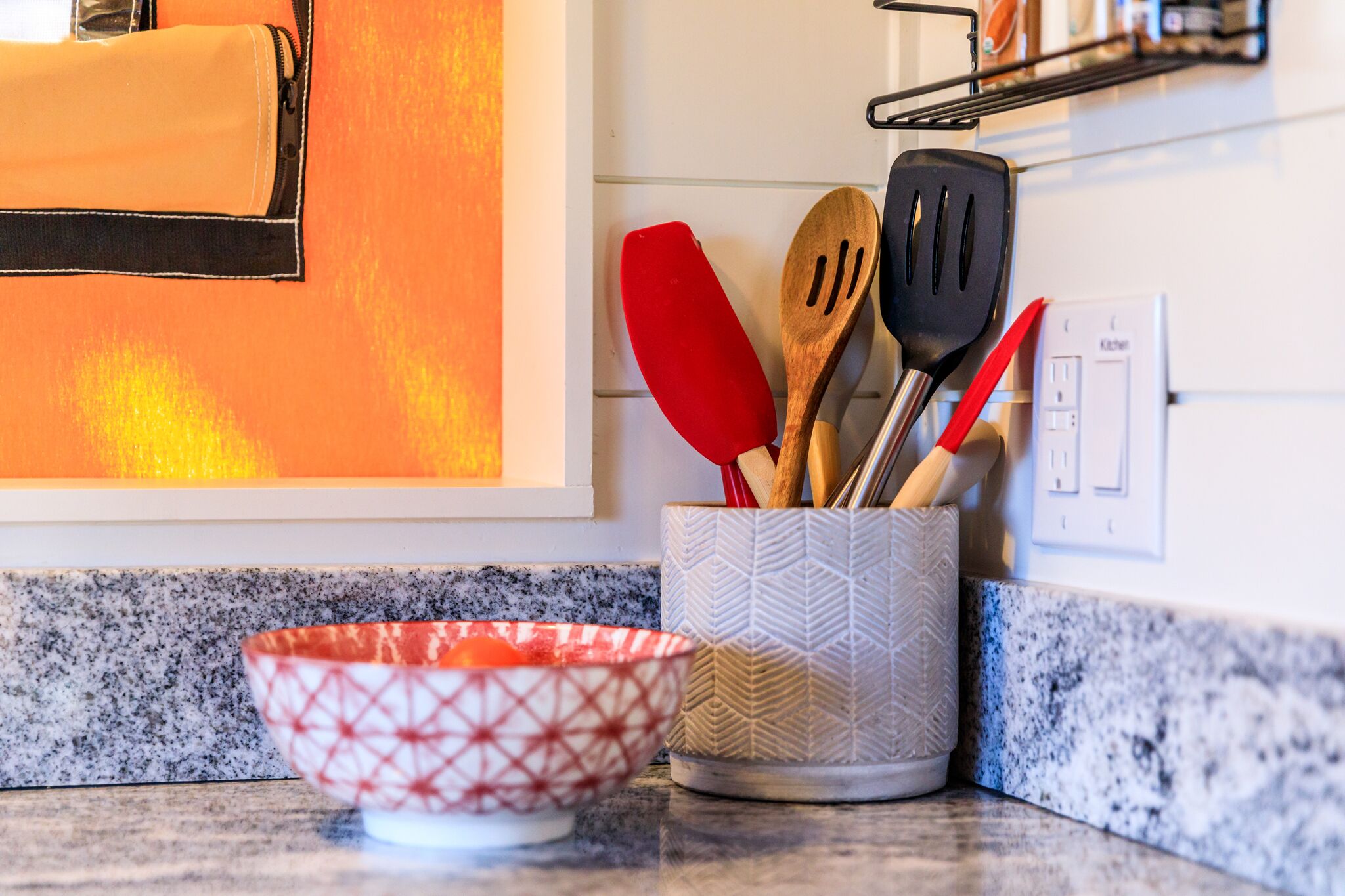 Una encimera de cocina con salpicadero de granito, un tazón blanco y rojo estampado y un recipiente para utensilios con utensilios de madera y plástico.