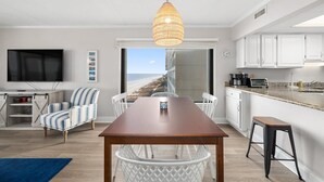 A modern kitchen with white cabinets, a dining area with a wooden table and chairs, a large window with a view of the beach, and a hanging light fixture. - Three Bedroom Direct Oceanfront Condo with Pool (Ocean City)