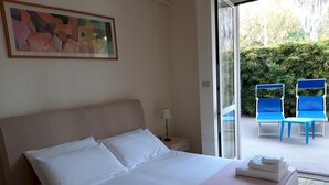 A hotel room with a bed, a nightstand, a lamp, and a window leading to an outdoor seating area with blue chairs. - Le Barchette 2 - Piano Terra (Lecce)