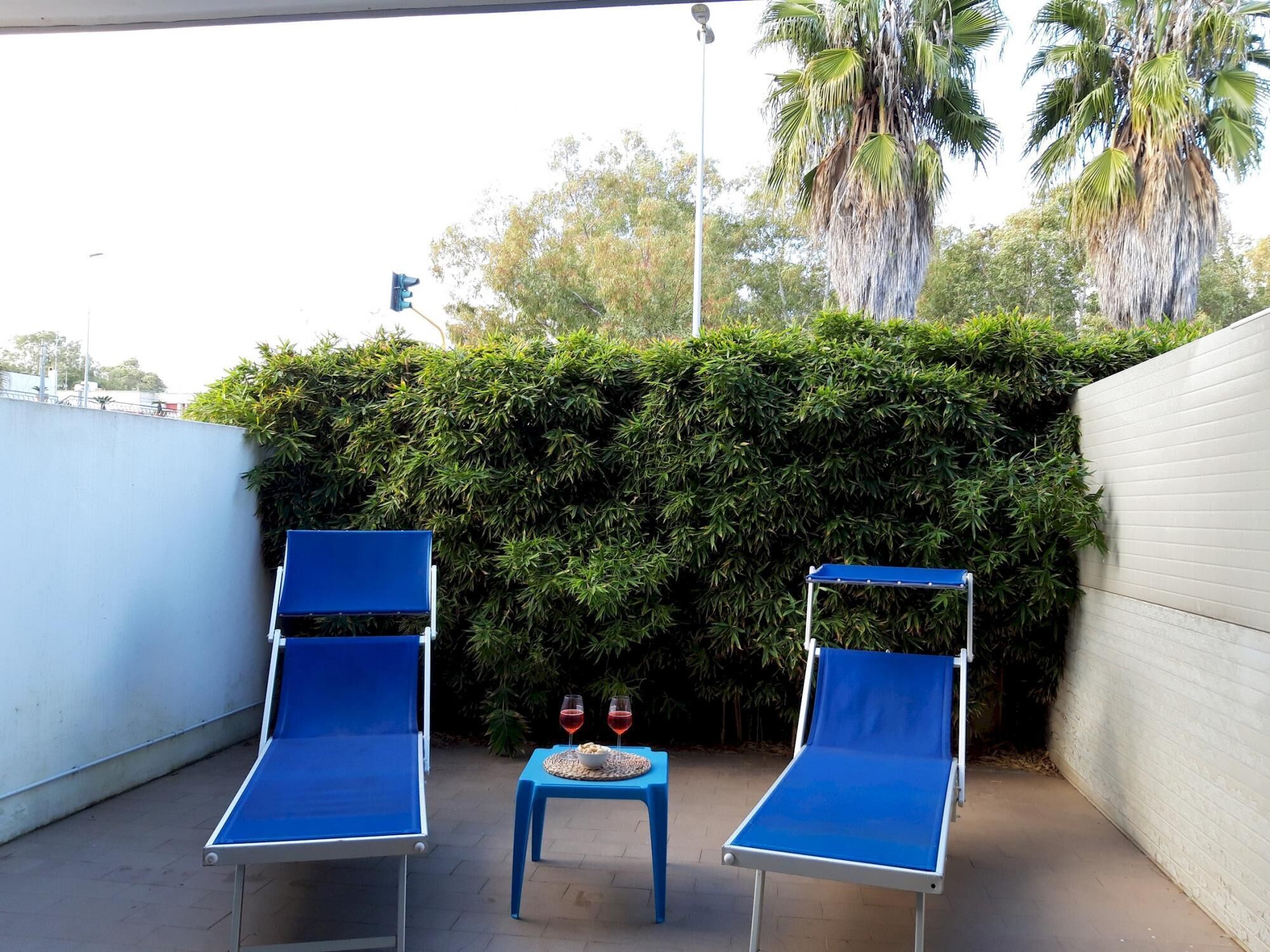 A patio with two blue lounge chairs, a small table with a bowl and two wine glasses, and a dense green hedge in the background.