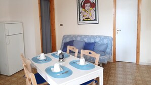 Un comedor con una mesa blanca preparada para cuatro personas, un sofá azul y un refrigerador.