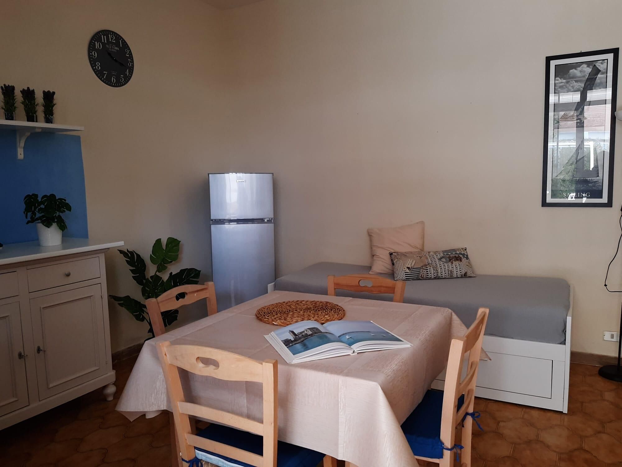A small, cozy room with a bed, a table with a book, a refrigerator, a clock, and a wall-mounted picture.