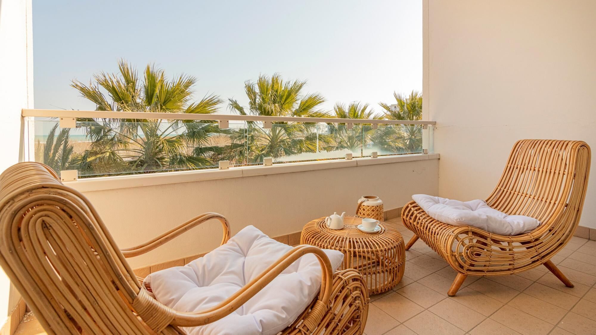 A balcony with wicker furniture, a table with a teapot and cup, and a view of palm trees.