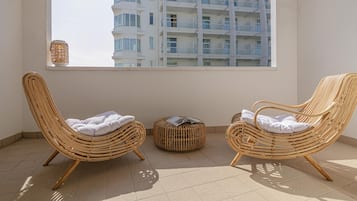 Two wicker lounge chairs with cushions, a small wicker ottoman, and a window with a view of a multi-story building.