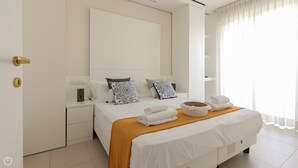 A modern hotel room with a large bed, white bedding, and decorative pillows. There is a wooden side table with a lamp and a small decorative item. The room has a large window with sheer curtains. - Residenza Excelsior 704 (Vasto)