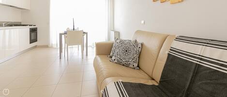 A modern living room with a sofa, a dining table, and a kitchen area with white cabinets and a stove.