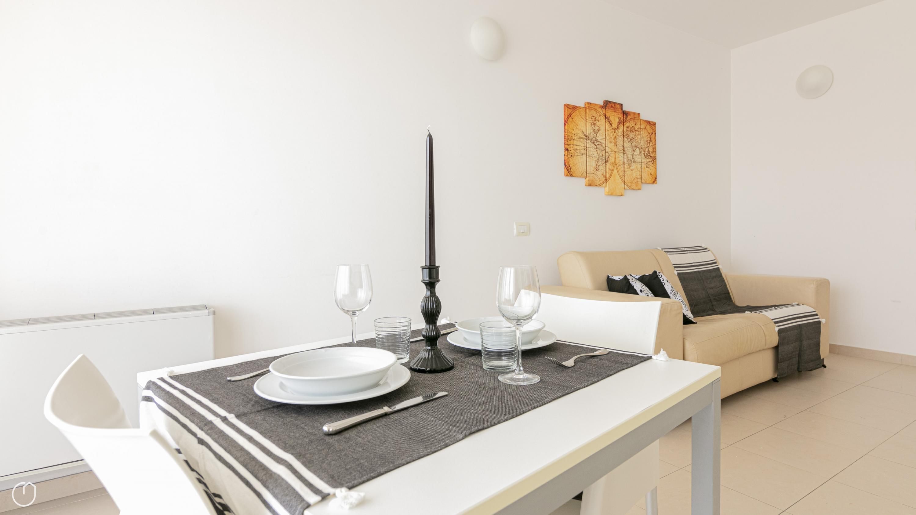 Taula de dinar preparada amb plats, gots i coberts, una espelma i un camavents gris. Al fons, un sofà beix amb una manta decorativa.