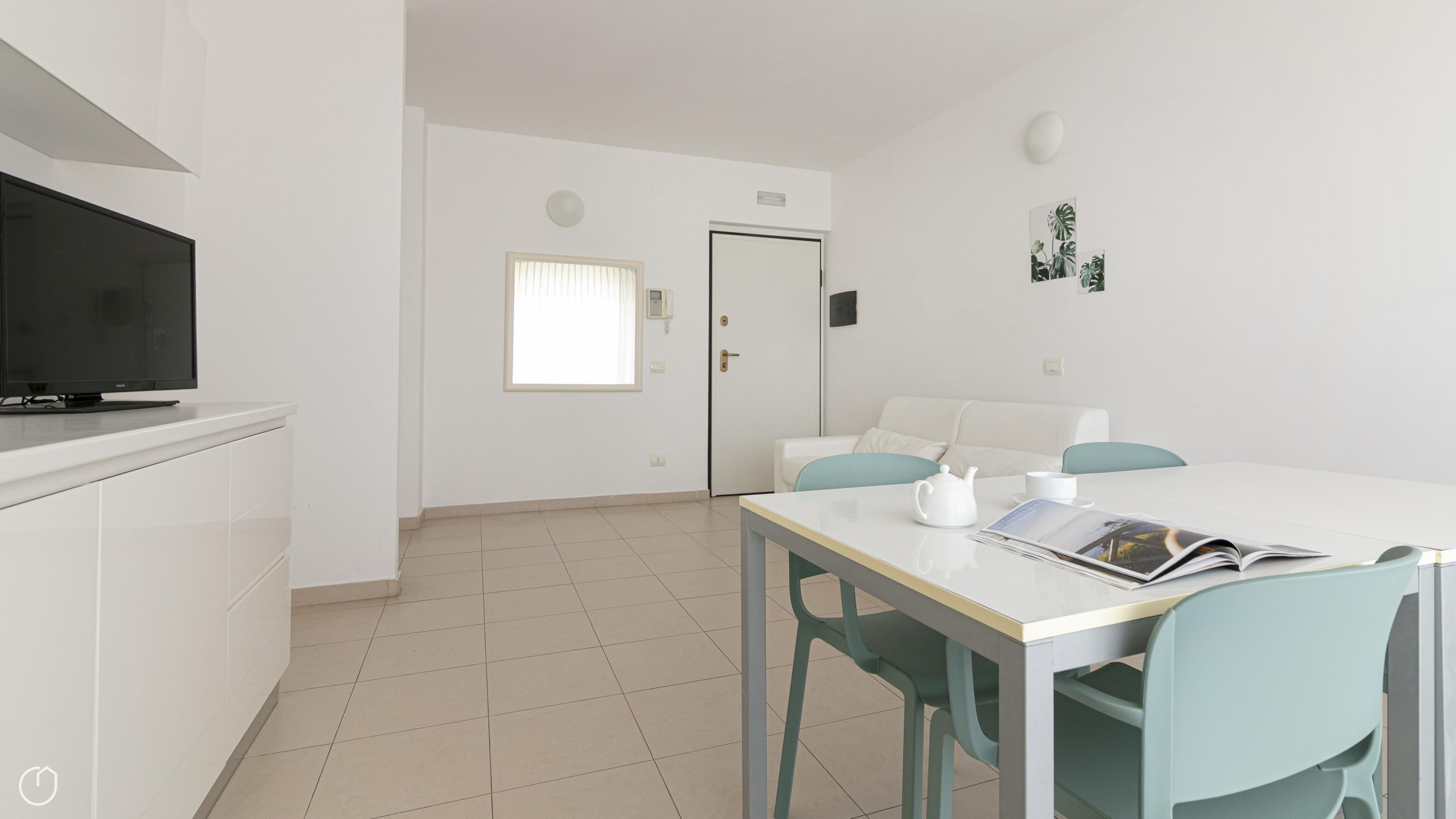 Ein modernes, minimalistisches Wohnzimmer mit Flachbildfernseher, Esstisch mit Stühlen, Sofa und einer Tür mit Fenster.