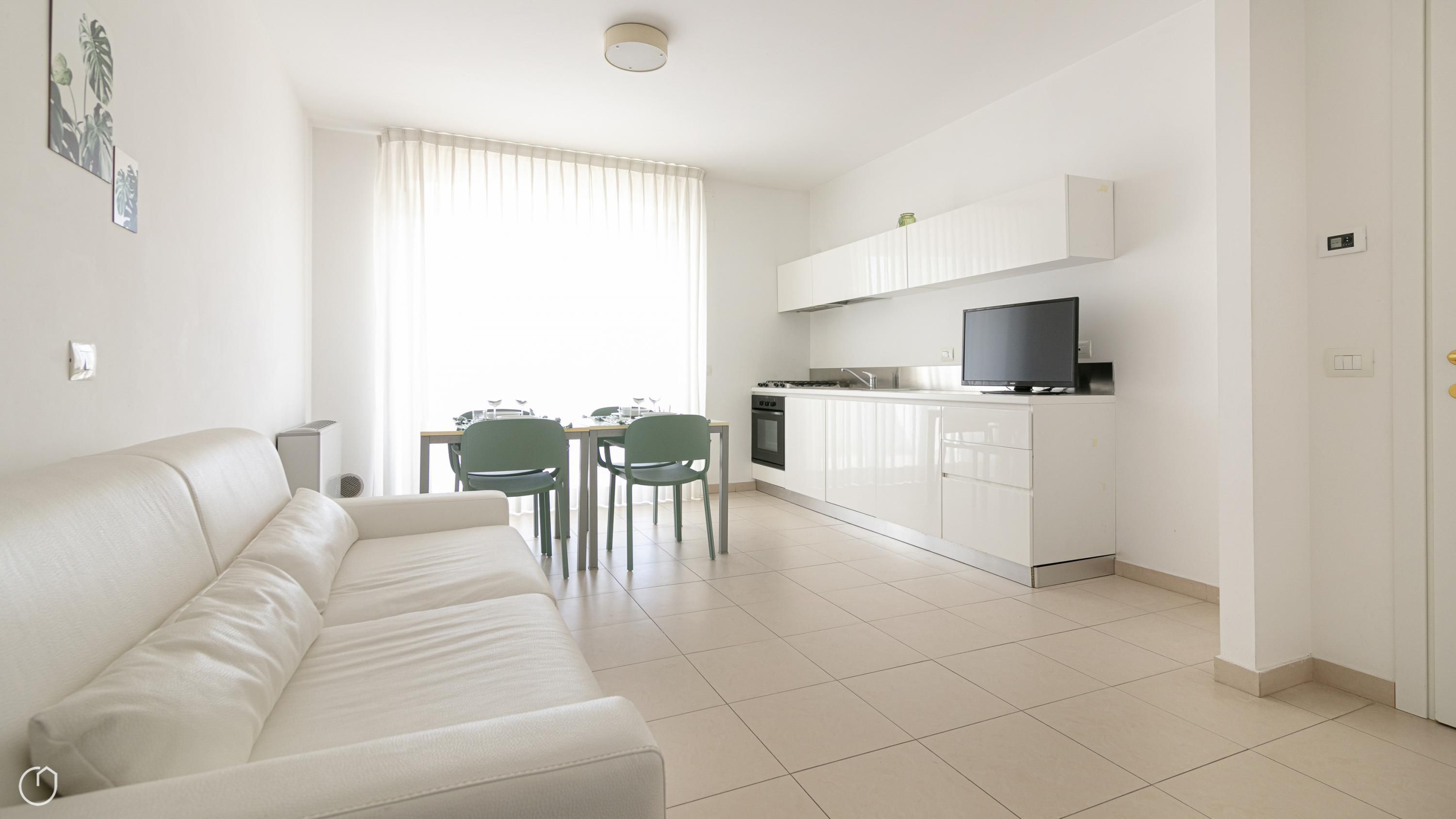 A modern living room with a white sofa, a dining area with two chairs, a kitchen with white cabinets and a TV on the counter.