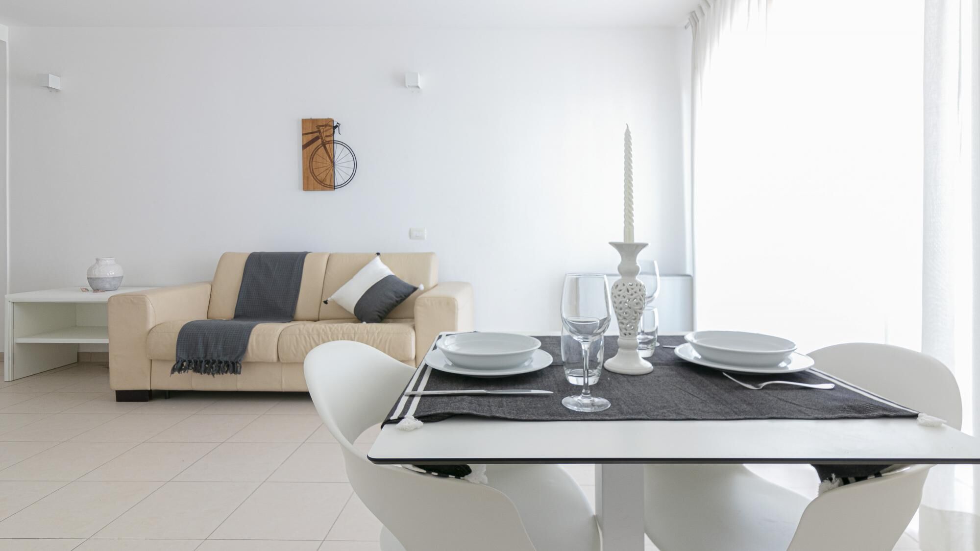 Una sala de estar moderna con un comedor. Incluye un sofá color beige con una manta gris, una mesa de centro blanca y una mesa de comedor dispuesta con platos, vasos y cubiertos. Hay un jarrón blanco sobre la mesa de centro y una obra de arte colgada en la pared sobre el sofá.