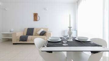 Una sala de estar moderna con un comedor. Incluye un sofá color beige con una manta gris, una mesa de centro blanca y una mesa de comedor dispuesta con platos, vasos y cubiertos. Hay un jarrón blanco sobre la mesa de centro y una obra de arte colgada en la pared sobre el sofá.