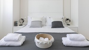 Una cama bien hecha con sábanas blancas, dos mesitas de noche con lámparas decorativas y una cesta de mimbre con toallas sobre una colcha gris con textura.