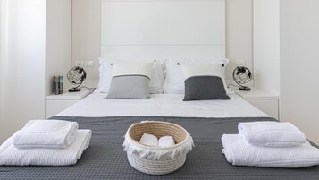 Un letto ben rifatto con lenzuola bianche, due comodini con lampade decorative e un cesto di vimini con asciugamani su una coperta grigia e texturizzata.