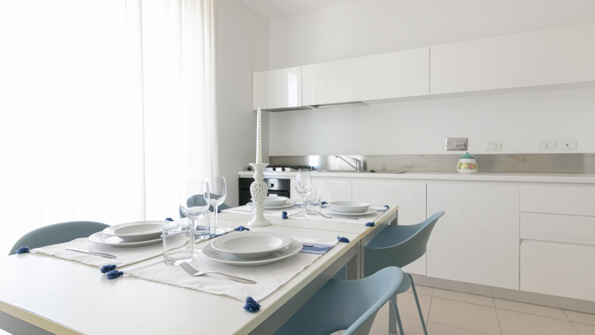 Una cocina moderna con armarios blancos, una mesa de comedor con platos, vasos y cubiertos, y sillas azules.
