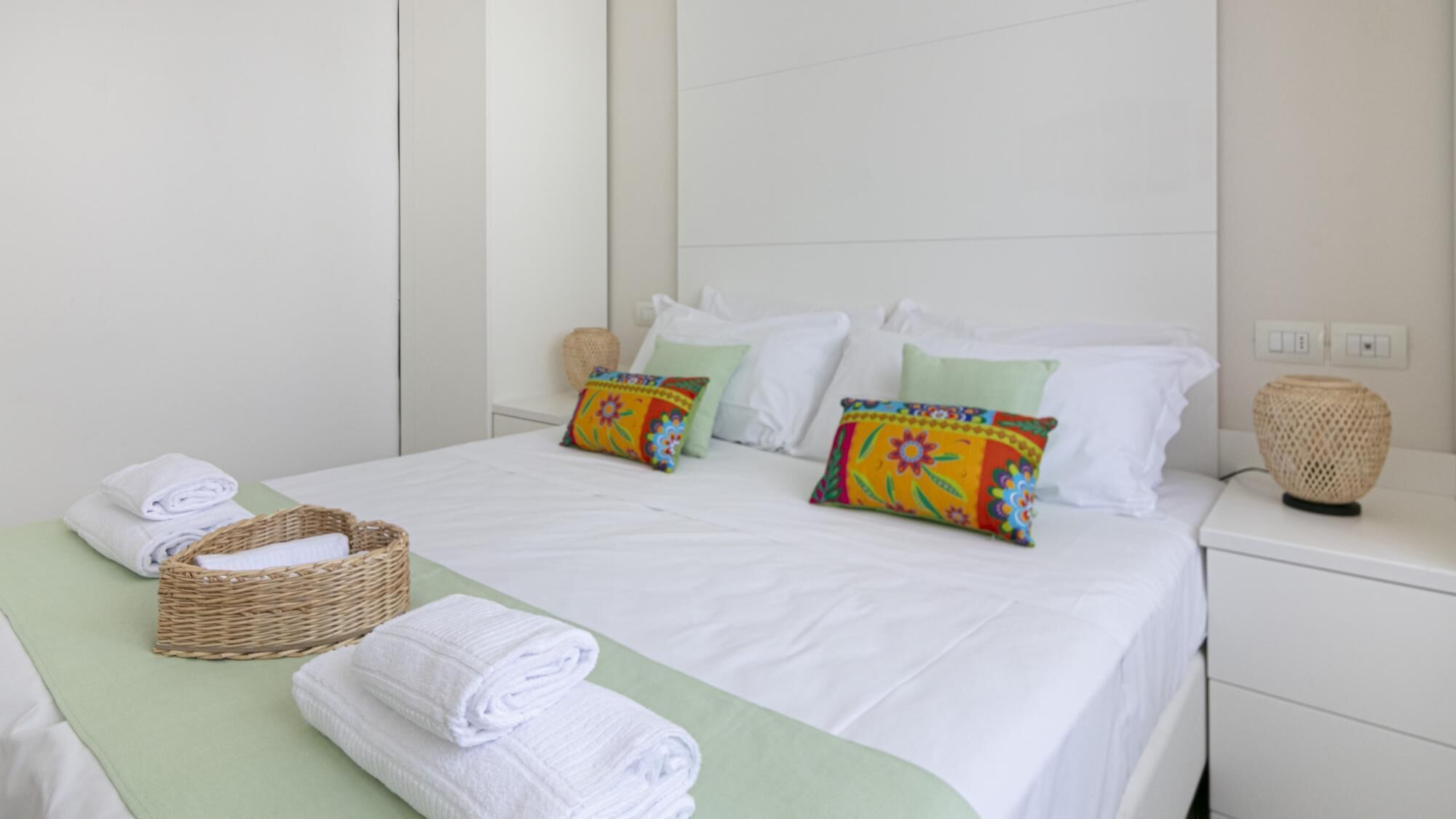 A neatly made bed with white linens, two colorful pillows, a wicker basket, and a bedside lamp on a white nightstand.